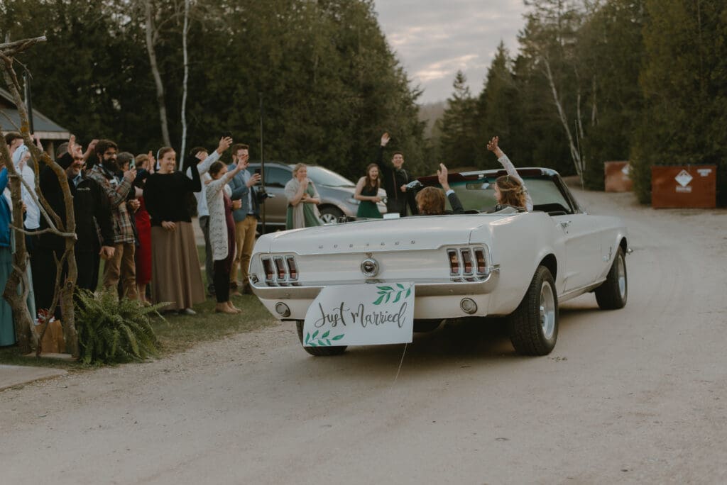 Bride and groom waving goodbye to guests as they drive away into forest in Markdale, Ontario at Campfire Bible Camp.