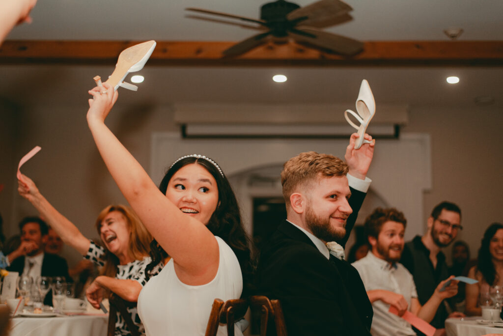 A bride and groom playing the shoe game at reception, raising bride's shoe, while the guests participate and guess in the background in Ottawa's Strathmere Wedding Estate.
