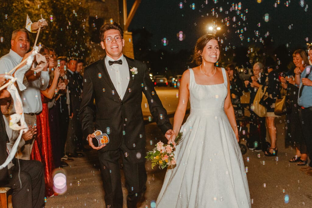 A bride and groom smiling and walking through their guests blowing bubbles at night as they exit their wedding reception in Waterloo, Ontario.