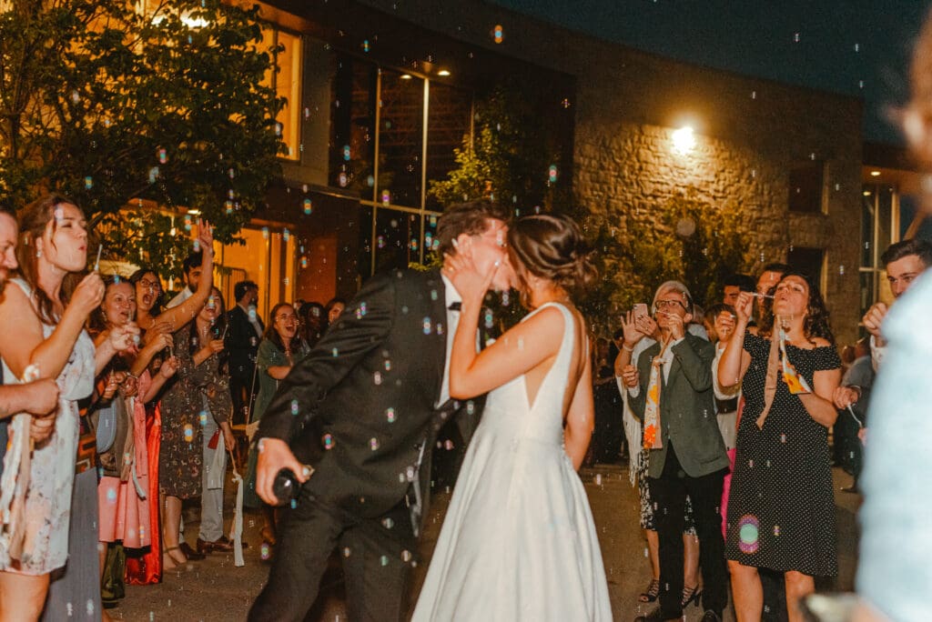 A bride and groom kissing and walking through their guests blowing bubbles at night as they exit their wedding reception in Waterloo, Ontario.