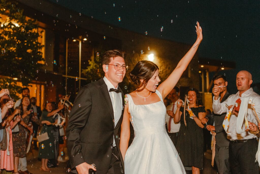 A bride and groom smiling, waving, and walking through their guests blowing bubbles at night as they exit their wedding reception in Waterloo, Ontario.