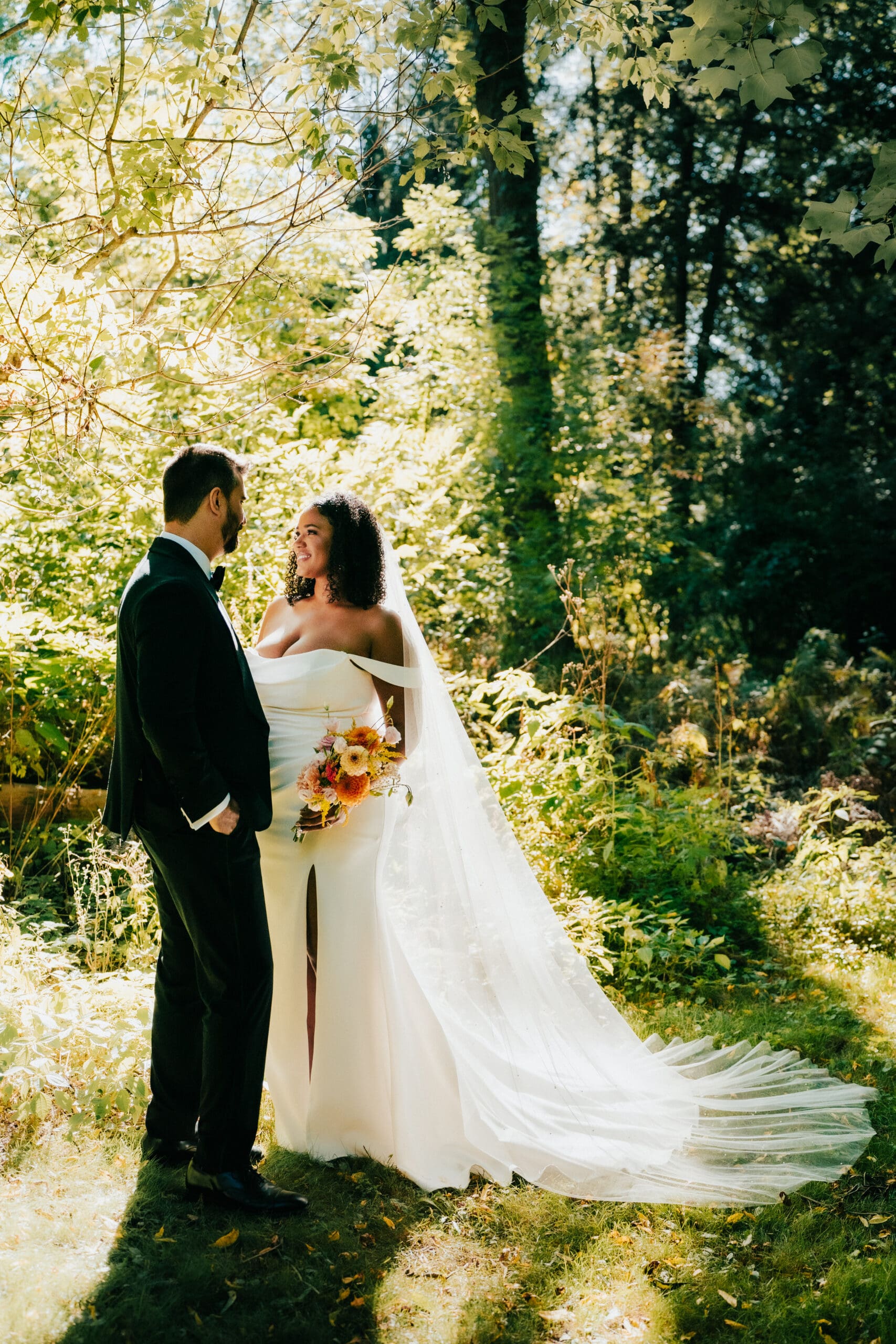 A bride and groom looking at each other lovingly holding flowers in the forest with sunlight in Hamilton Ontario at Fieldcote Memorial