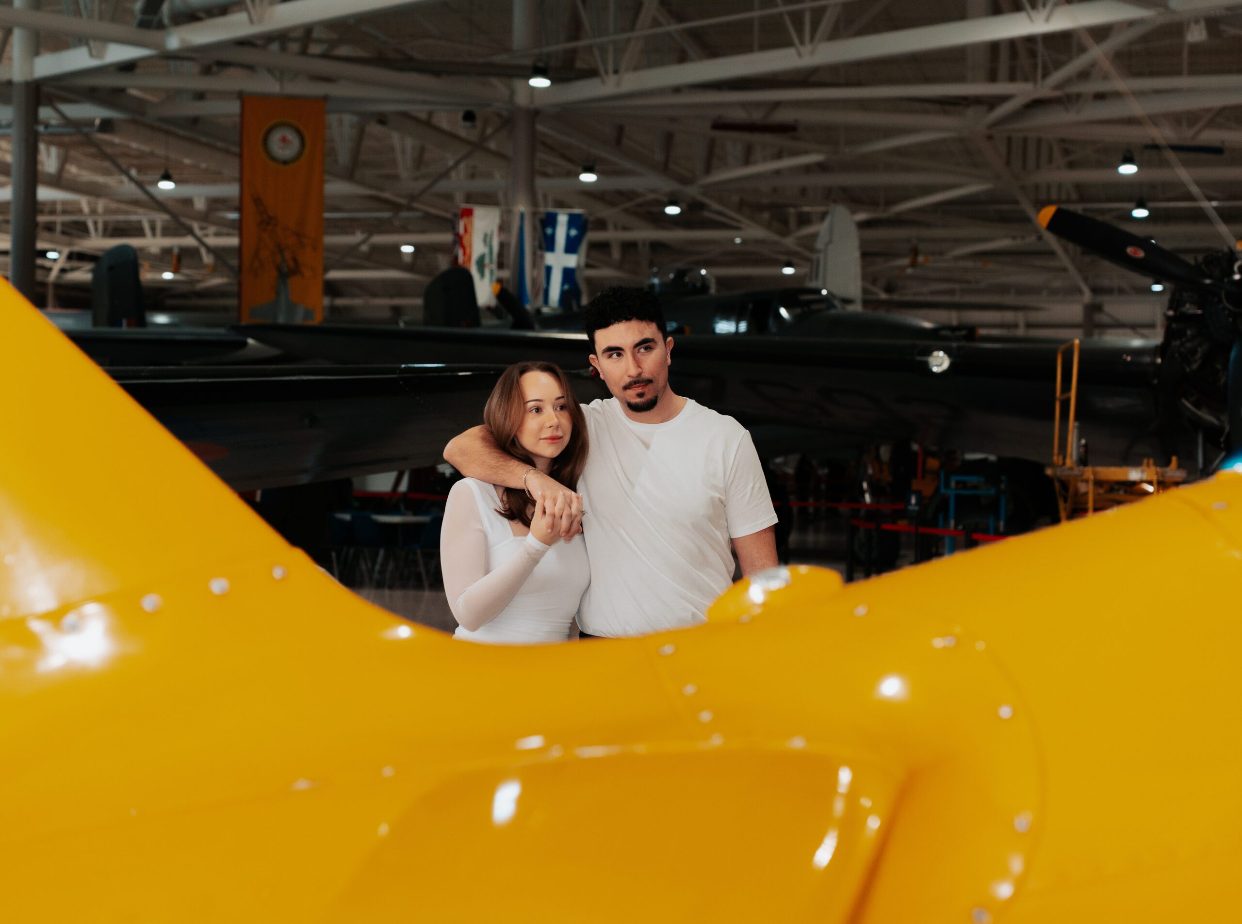 Man wrapping arm around girl as they look off into the distance, with a yellow plane in front at the Hamilton Warplane Museum at an engagement shoot.