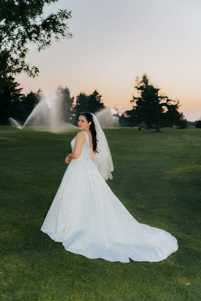 Editorial back-profile of bride TJ McGibbon in a satin wedding gown with dramatic water spray from golf course sprinklers at Buncrana Golf Club, Hamilton.