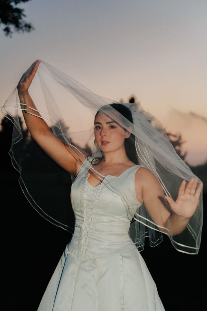Editorial close-up portrait of bride TJ McGibbon holding her veil up against a sunset sky, Hamilton wedding photography.