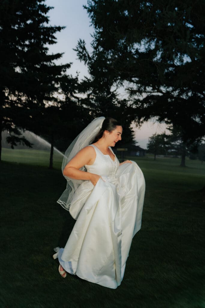 Bride TJ McGibbon laughing and running through grass holding her wedding dress as sprinklers spray behind her, documentary wedding style.
