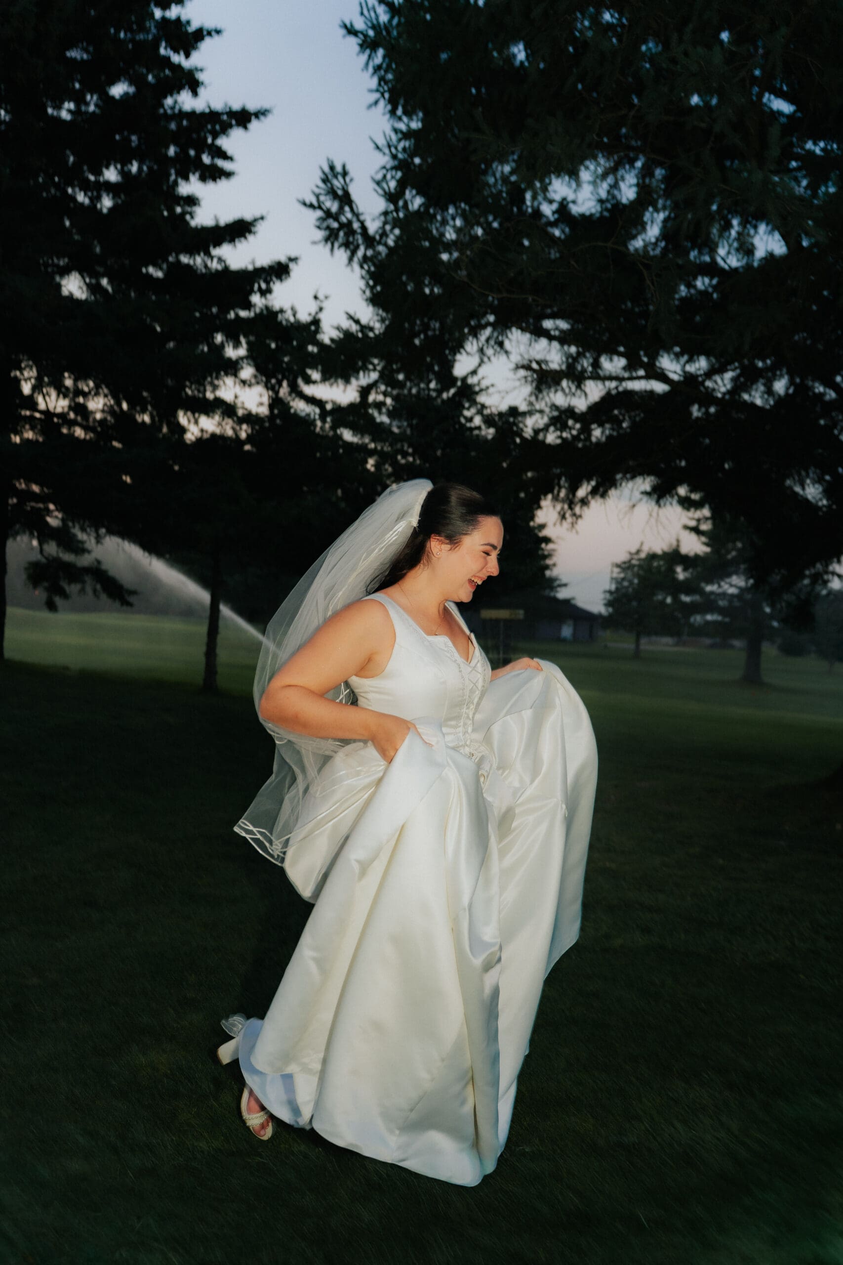 Bride smiling and holding her dress under a tree on the grounds of Buncrana Golf Course in Hamilton Ontario, natural light outdoor wedding portrait.