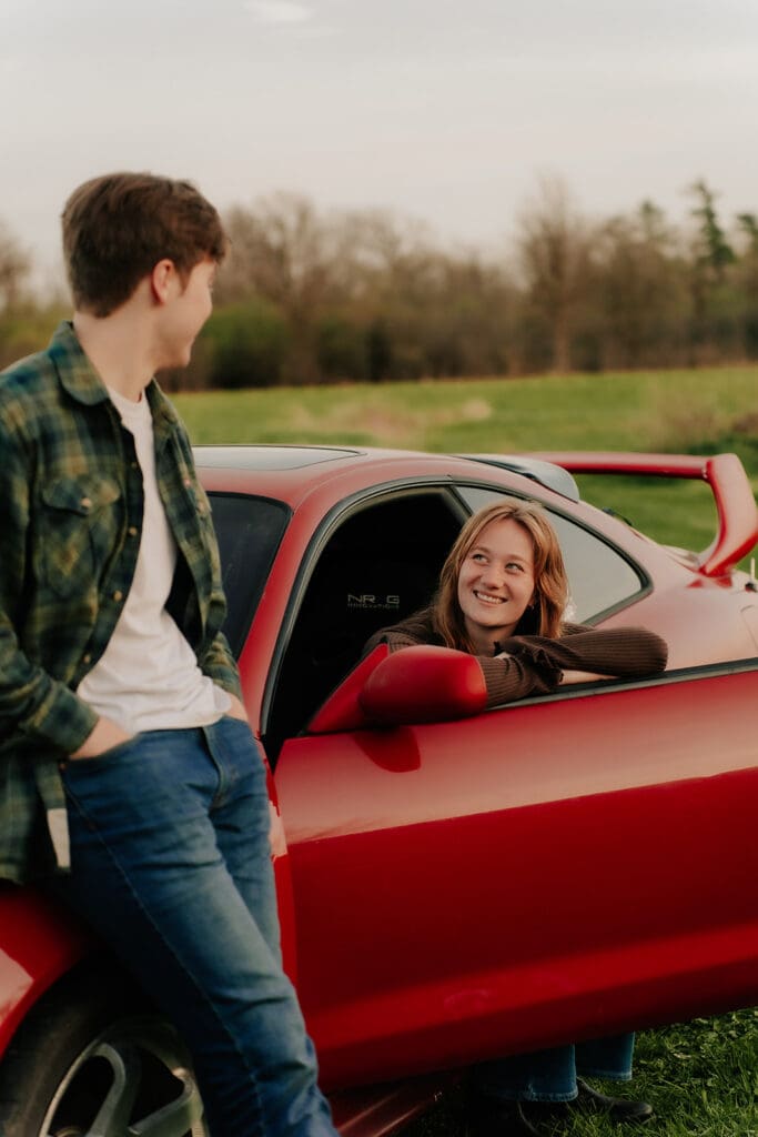 Girl in red car smiling at boy leaning on car. Hamilton Wedding Photographer